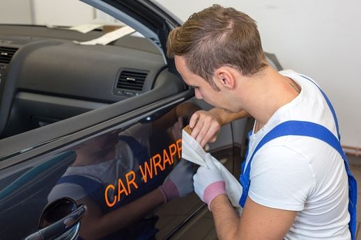 Mann in weißem T-Shirt und blauer Latzhose bringt Schriftzug 'CAR WRAP' auf schwarze Autotür an